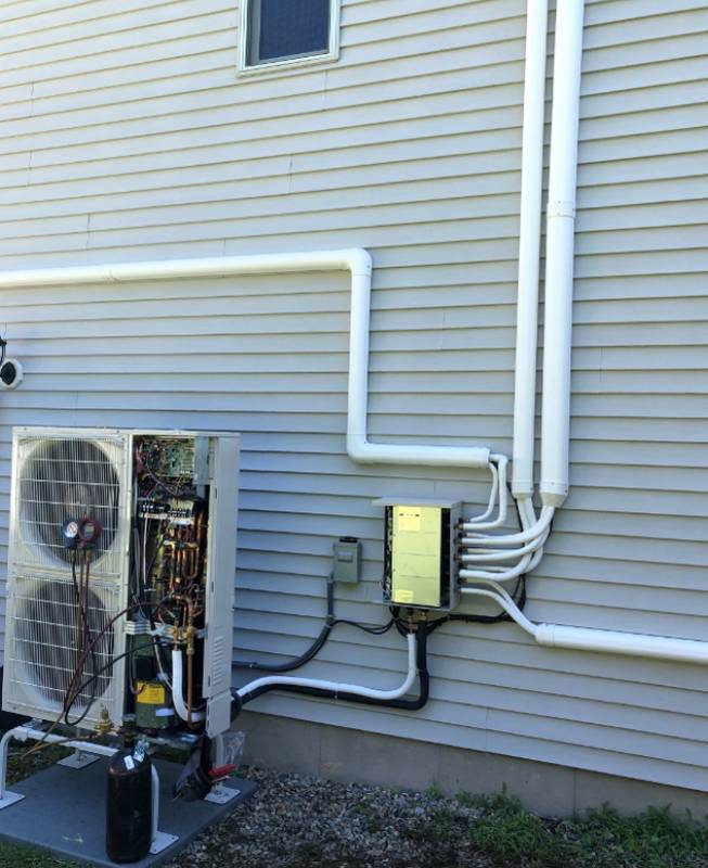 Mini-split outdoor unit on vinyl siding with its panel open and gauges attached, white line-set covers running up the wall