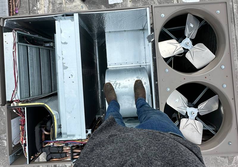 Overhead view of a large packaged unit with its panels off and two condenser fans, a technician standing inside the blower section