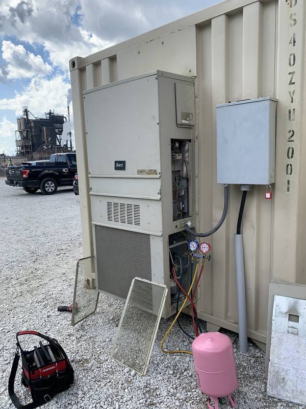 Wall-mount AC unit on a shipping container with its panel open and a gauge manifold hooked up, a refrigerant tank and tool bag on the gravel